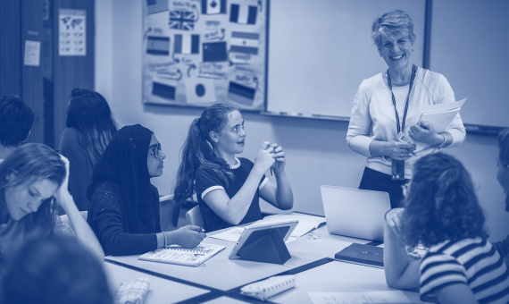 Female teacher standing and smiling in a classroom in front of a table of female pupils who are working.