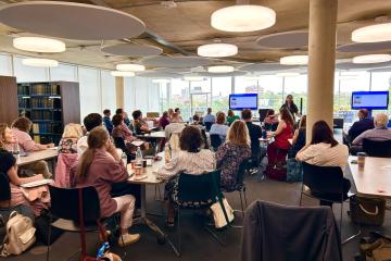 Conference scene from 2025 of the back of peoples' heads looking towards a speaker in a modern room