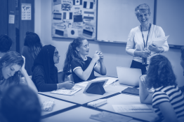 Female teacher standing and smiling in a classroom in front of a table of female pupils who are working.