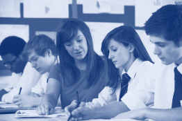 Female teacher sitting at a table with secondary school students, she is helping a female student with some written work.