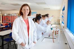 Lady stood smiling in a science lesson wearing a white lab coat and safety goggles