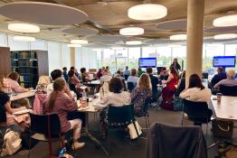 Conference scene from 2025 of the back of peoples' heads looking towards a speaker in a modern room